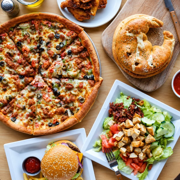 Overhead spread of loaded pizza, calzone, chicken wings, cheeseburger with fries, and grilled chicken salad on a wooden table.
